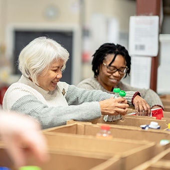 A small group of four volunteers are seen standing at a table full of cardboard boxes as they pack them with non-perishable food items at a local Food Bank.  A young mixed race girl is working with three seniors as they happily give of their time for  the cause and carefully pack the items into the boxes.
