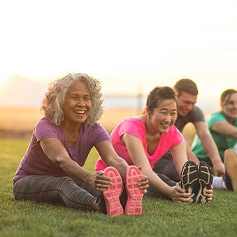 A group of adults attending a fitness class outdoors are doing leg stretches. The participants are arranged in a line. The focus is on a mature woman who is smiling toward the camera.