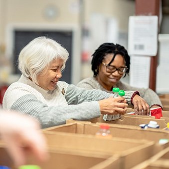 A small group of four volunteers are seen standing at a table full of cardboard boxes as they pack them with non-perishable food items at a local Food Bank.  A young mixed race girl is working with three seniors as they happily give of their time for  the cause and carefully pack the items into the boxes.