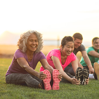 A group of adults attending a fitness class outdoors are doing leg stretches. The participants are arranged in a line. The focus is on a mature woman who is smiling toward the camera.