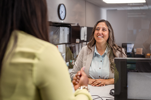A friendly female customer service representative of Latin American descent greets a customer with a big smile at the counter in a bank.