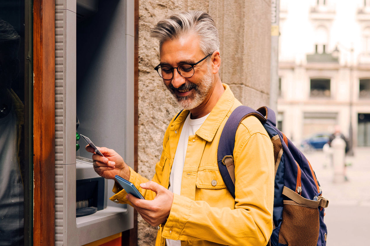 Tourist holding a smartphone and debit card stands in front of an ATM. 