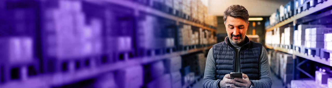 Man standing in warehouse aisle entering data into a smartphone. 